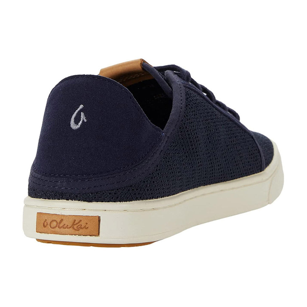 Navy blue OLUKAI PEHUEA LI TRENCH BLUE sneaker with a white, non-marking rubber outsole and a wooden heel label bearing the brand name &quot;Olukai&quot;, shown from a rear side angle.