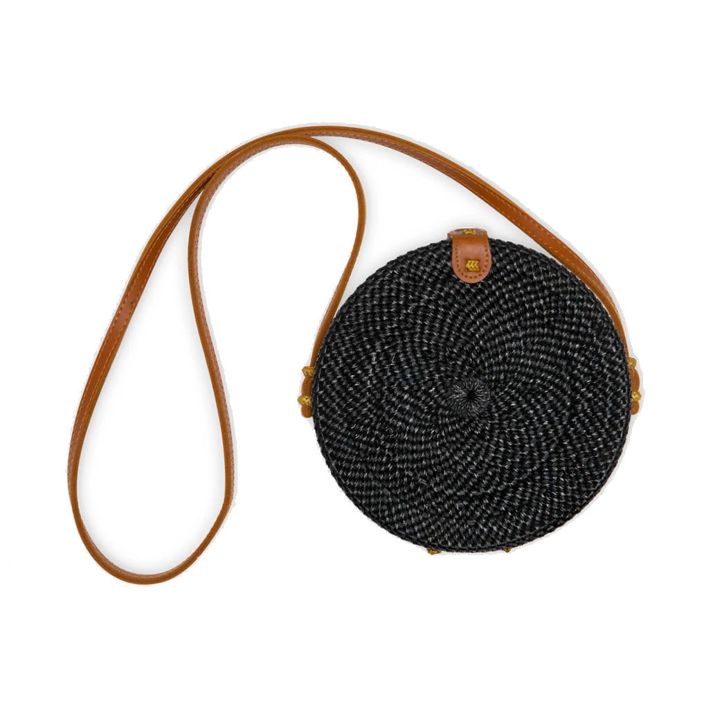 Round black POKOLOKO Circle Rattan Bali Bag with a long, brown vegan leather strap and a small vegan leather flap closure, displayed on a white background.
