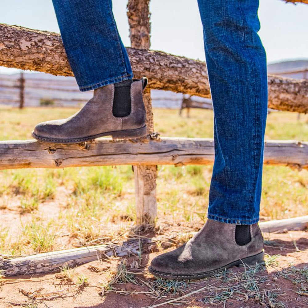 A person wearing Born Shane Chelsea Boot Suede Taupe boots and blue jeans stands with one foot on a wooden fence rail in a dry grassy area.