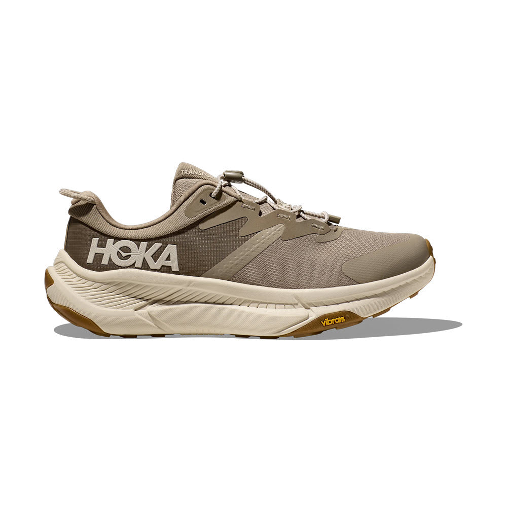 A single Hoka TRANSPORT DUNE/EGGNOG - MENS trail running shoe in beige and white colors, viewed from the side, featuring a thick, cushioned sole made from eco-friendly materials and the brand logo.