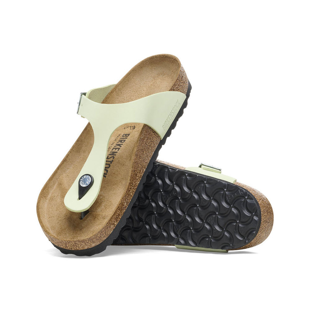 A pair of light green nubuck leather Birkenstock Gizeh sandals with cork-latex footbeds and black soles, arranged at a tilted angle against a white background.