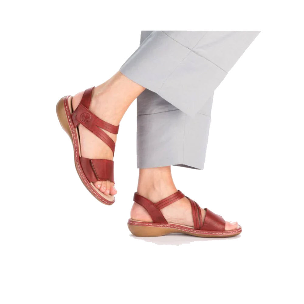 Woman&#39;s feet walking, wearing RIEKER COMFORT BOTTOM ASYMMETRICAL SANDAL RED - WOMENS sandals and light gray cropped trousers, shown against a white background.