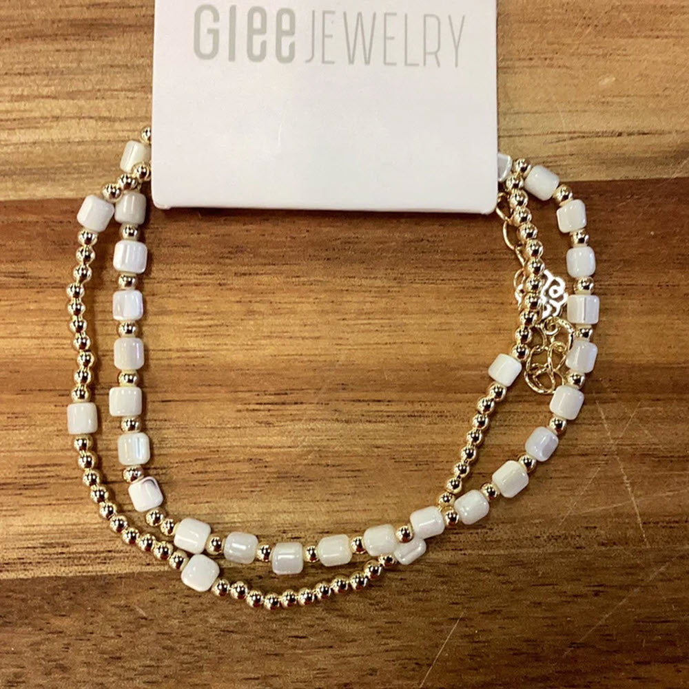 A double-stranded white beaded necklace with gold accents displayed on a wooden surface, featuring Glee jewelry and labeled &quot;GLEE&quot; above it.