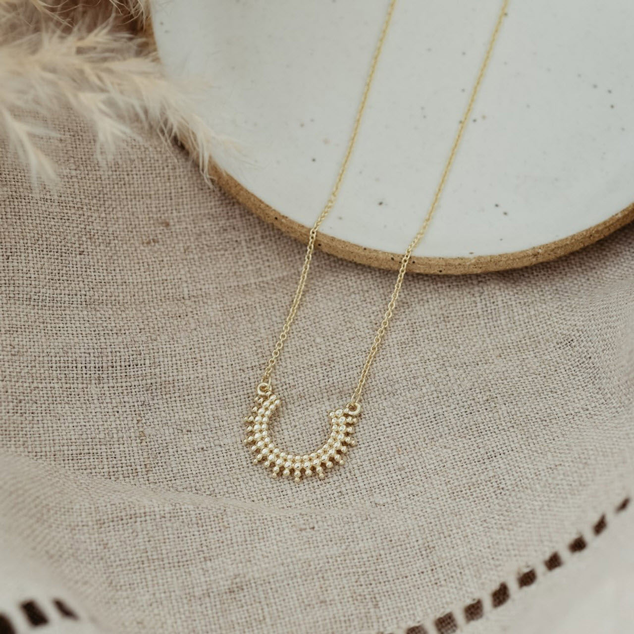 The Glee Curved Luck Necklace Gold displayed on a beige fabric surface, with a ceramic plate in the background. This nickel-free jewelry piece ensures hypoallergenic qualities for sensitive skin.