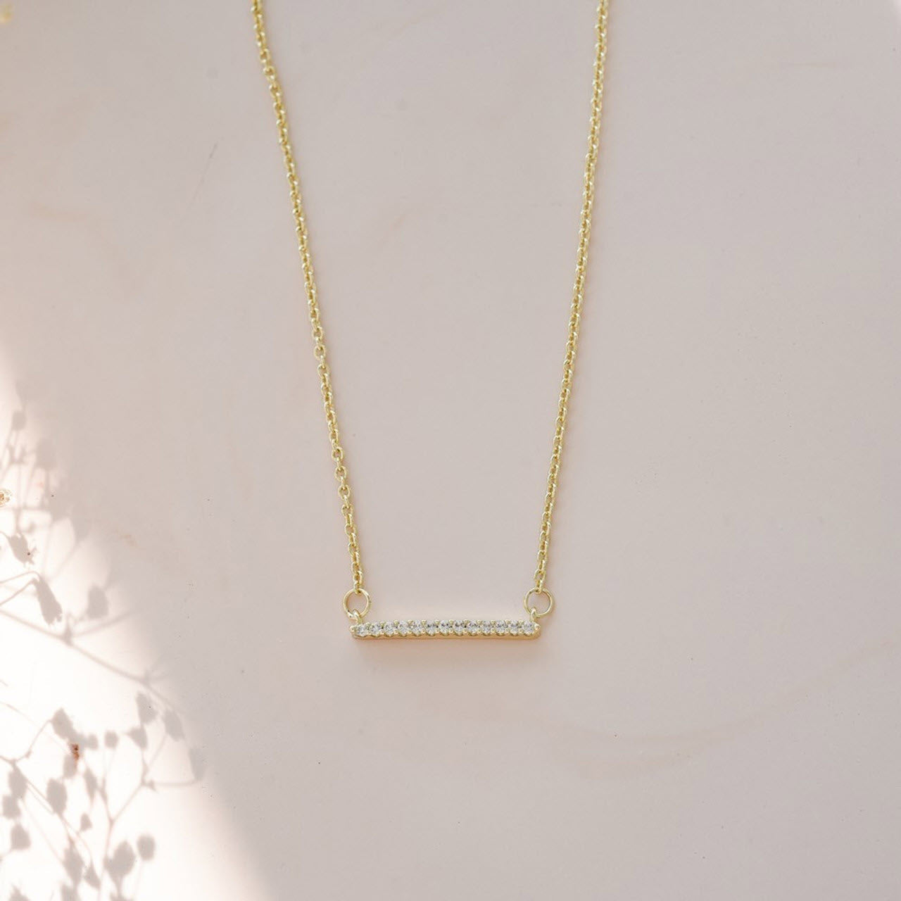 A delicate Glee gold necklace with an anti-tarnish horizontal bar pendant adorned with small crystals, displayed on a soft, shadowed background.
