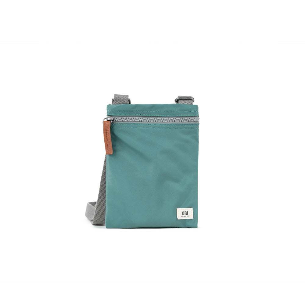 ORI LONDON CHELSEA CROSSBODY SAGE from the Ori London collection with a zipper and a brand logo, featuring a grey strap and a brown leather pull tab, on a white background.
