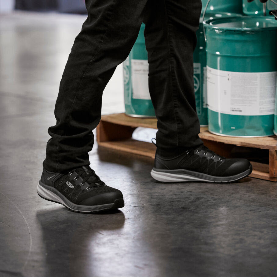 Person in Keen Composite Toe Vista Energy Vapor/Black - Mens work shoes standing next to hazardous material containers in an industrial setting.