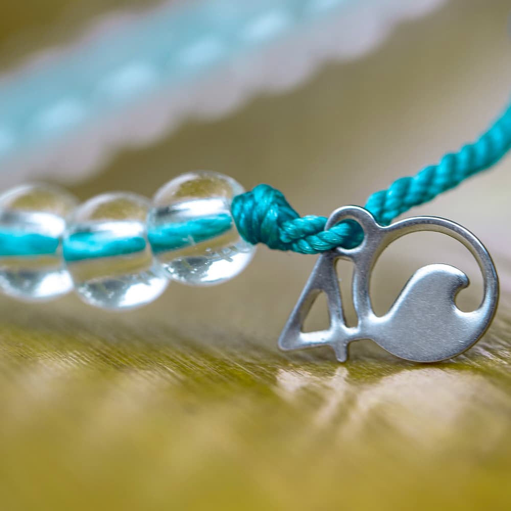 A close-up of a 4Ocean ocean conservation bracelet with translucent beads and a silver charm in the shape of a dolphin on a textured golden surface.