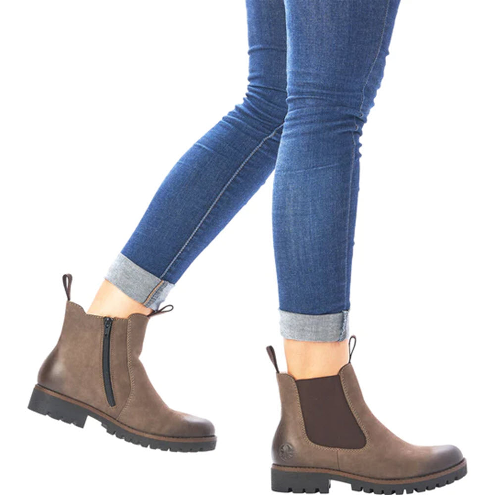 A person wearing blue jeans and RIEKER LUG SOLE CHELSEA WITH ZIP MUD, a women&#39;s leather ankle boot by Rieker, walks against a white background.
