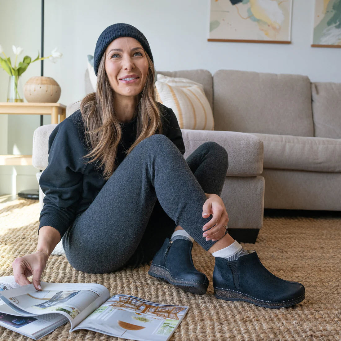 Woman in casual attire and Dansko Makara booties sitting on the floor of a living room, reading a magazine, with a smile on her face and maps on the wall behind.