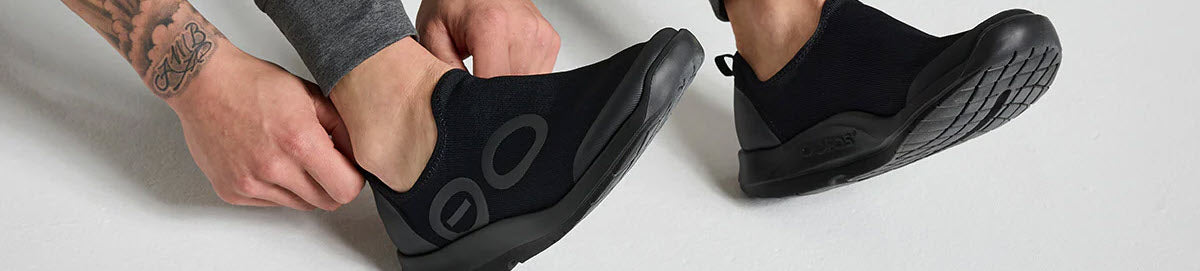 Person wearing black slip-on shoes with a unique sole design, adjusting one shoe with their hands.