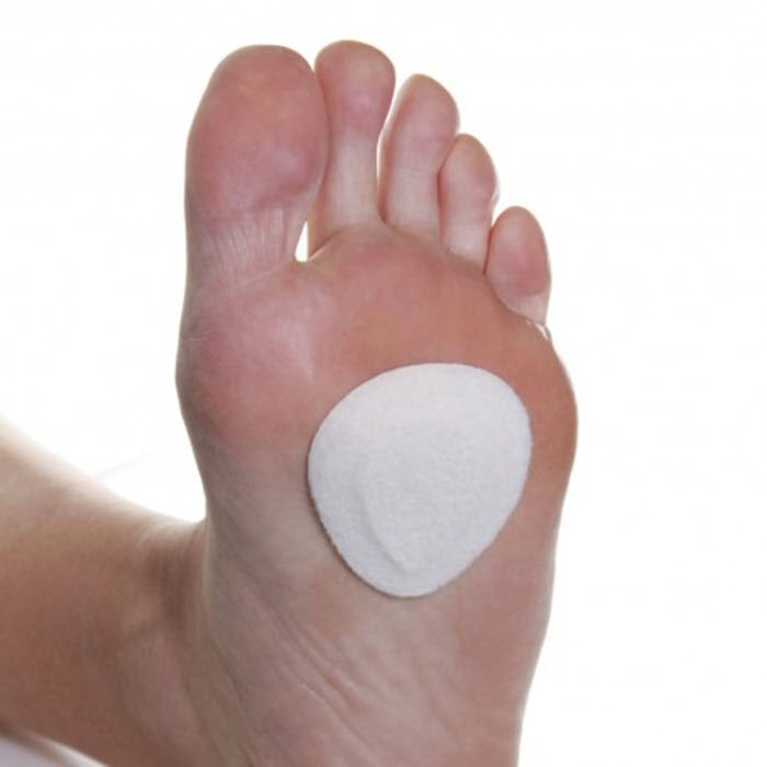 A close-up of a human foot with a round white Dr. Jill&#39;s LW FOOT CARE ADHESIVE METATARSAL PAD applied to the sole, isolated on a white background.