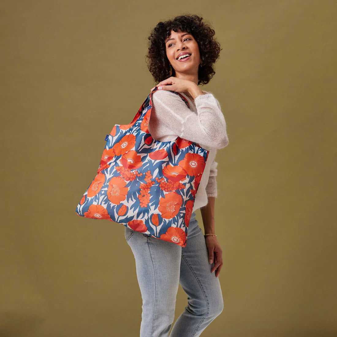 A woman with curly hair, smiling, holding a large eco-friendly Rockflowerpaper BLU BAG ICELANDIC POPPIES, wearing a beige sweater and jeans against a tan background.