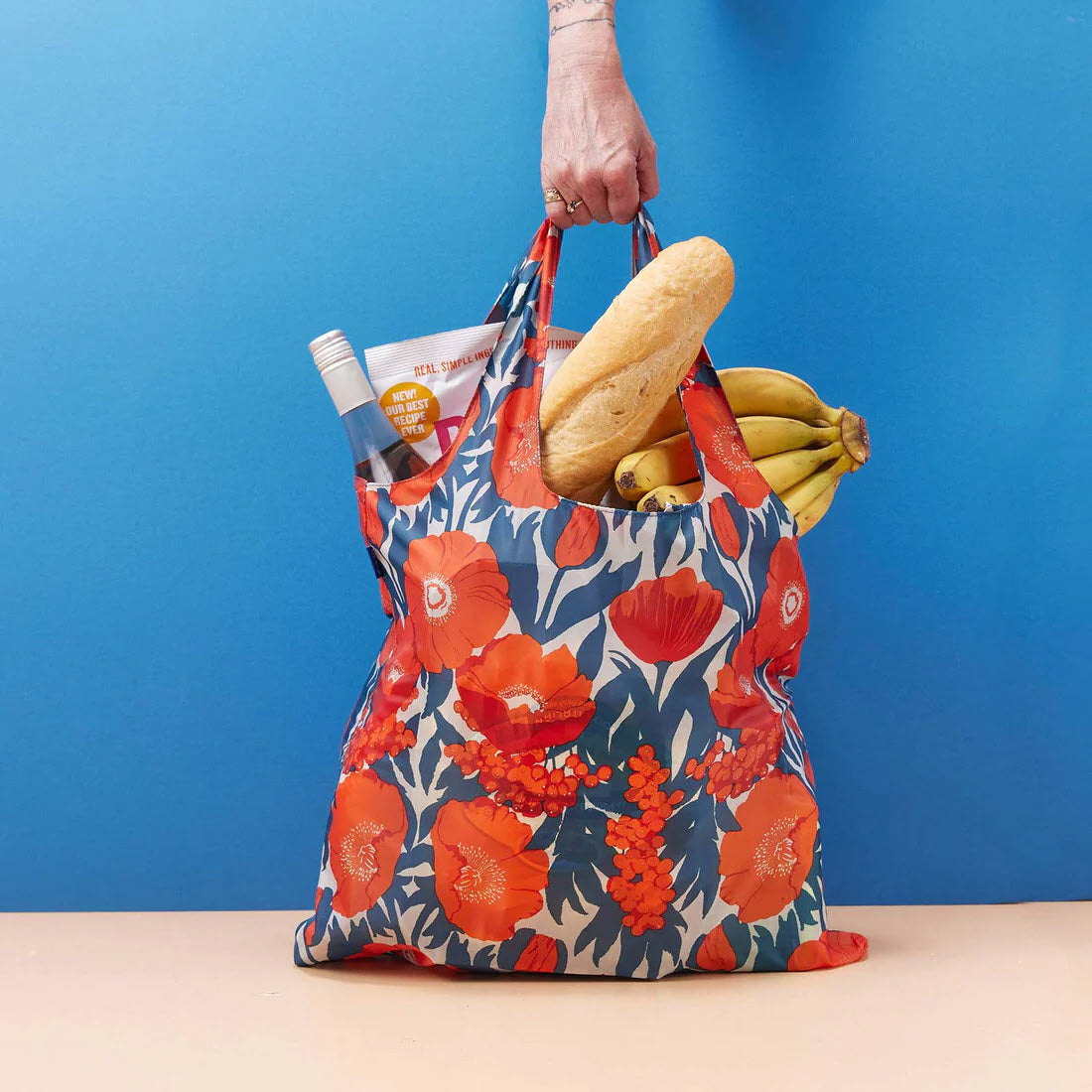 A person holding a Rockflowerpaper BLU BAG ICELANDIC POPPIES filled with groceries including a baguette and bananas against a blue background.