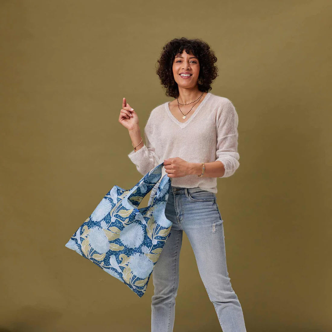 A woman with curly hair, smiling and pointing upwards, holding a colorful Rockflowerpaper BLU BAG CHRYSANTHEMUM tote bag, dressed in a beige sweater and ripped jeans against a tan background.