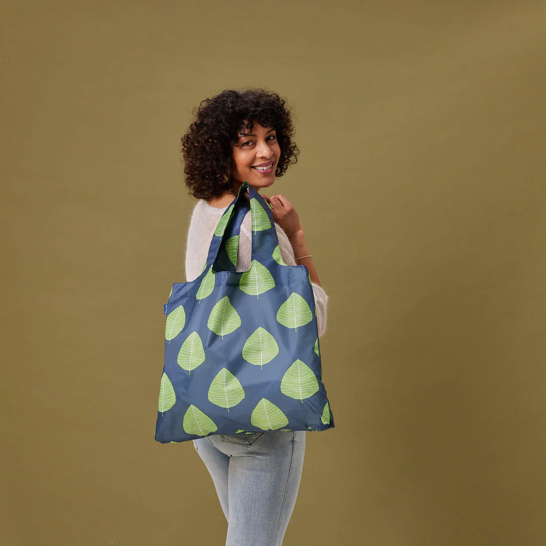 A smiling woman with curly hair holding a Rockflowerpaper BLU BAG ASPEN LEAVES stands against a beige background.