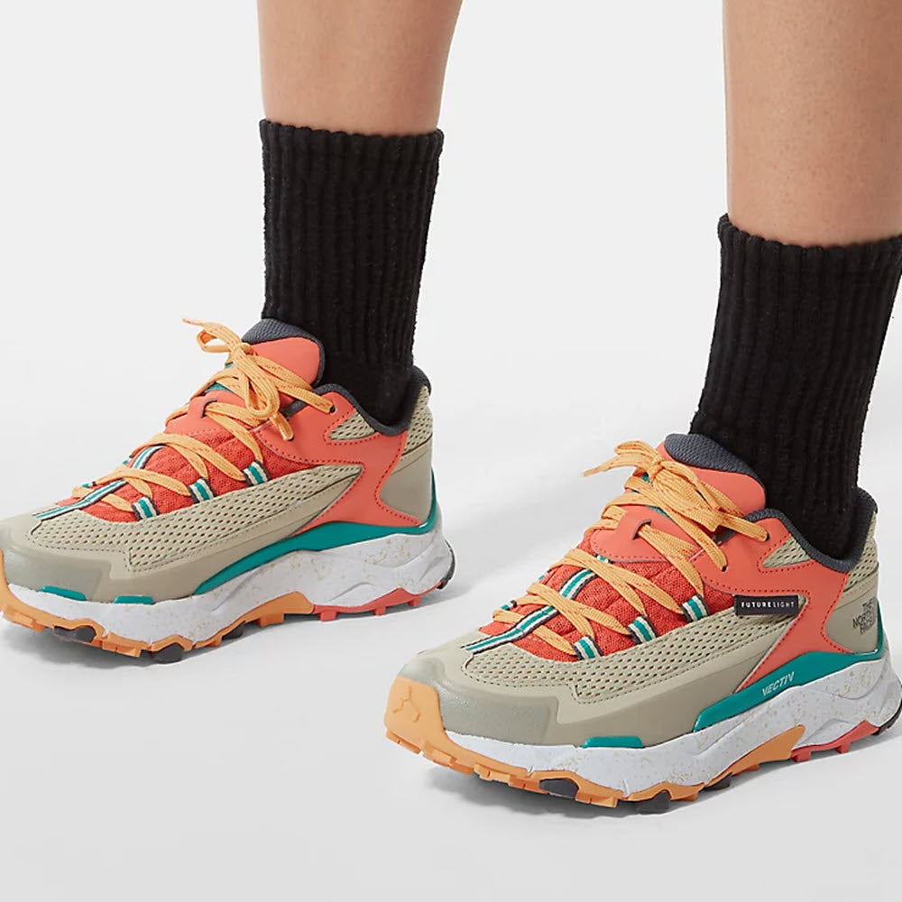 Person wearing colorful sneakers with THE NORTH FACE VECTIV TARAVAL FUTURELIGHT GRAVEL/CORAL SUNRISE - WOMENS technology paired with black socks, standing on a white surface.