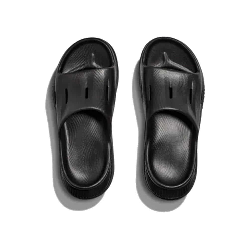 A pair of Hoka Ora Recovery Slide 3 black rubber slide sandals with a textured design and a sustainable sugarcane footbed, viewed from above on a white background.