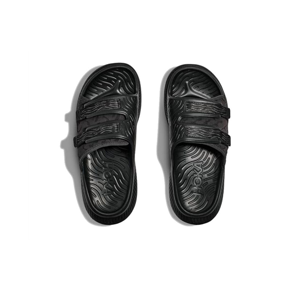 A pair of ORA LUXE BLACK/BLACK - ADULT sandals with textured insoles and adjustable Ariaprene® straps, photographed from above on a white background.