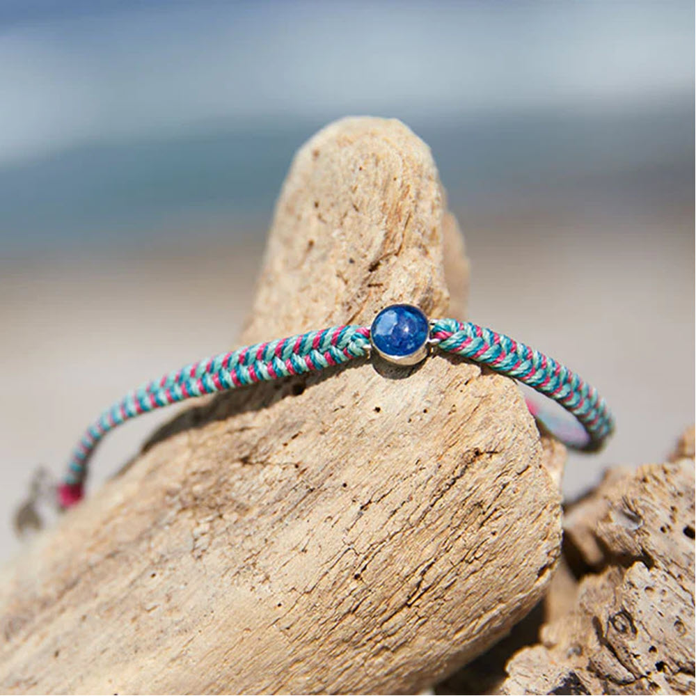 A vibrant &quot;4Ocean Ocean Drop Bracelet Tropical&quot; made from 4Ocean Plastic with a blue gemstone resting on weathered driftwood against a beach backdrop aims to raise awareness about microplastic pollution.