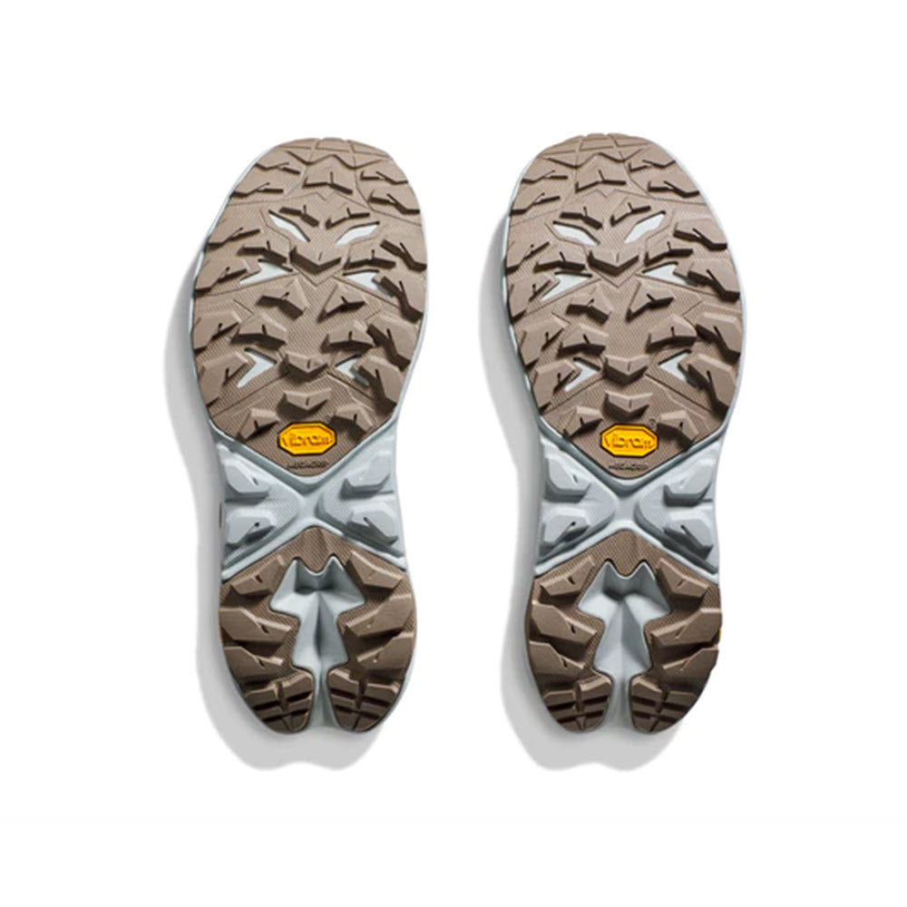 Two HOKA ANACAPA 2 MID GTX DUNE/ICE FLOW - WOMENS hiking boot soles placed side by side on a white background, featuring intricate Vibram Megagrip tread patterns in gray and brown colors.