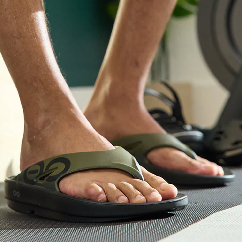 Close-up of a person&#39;s feet in OOFOS OOriginal Sport Tactical Green - Mens sandals standing on a gray exercise mat, with gym equipment in the background.