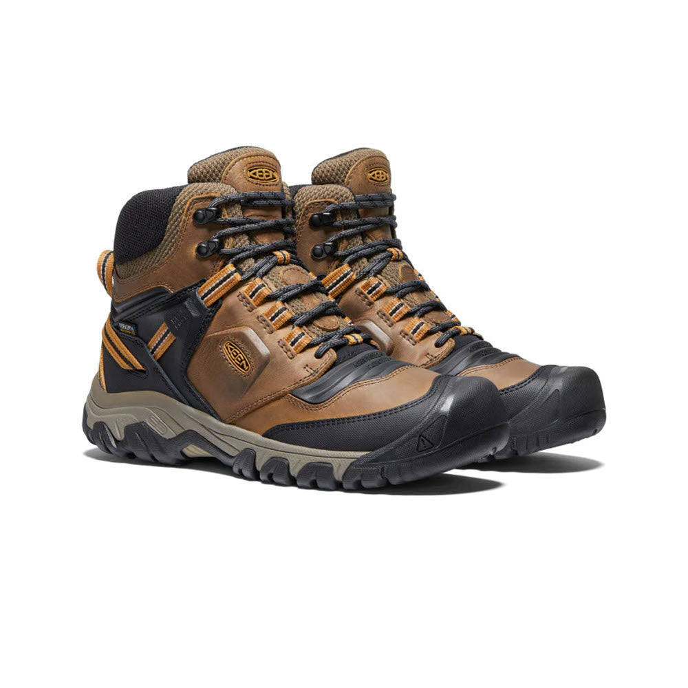 A pair of KEEN RIDGE FLEX MID WP BISON/GOLDEN BROWN - MENS high-top hiking boots with brown leather and black rubber soles, featuring orange accents and metal eyelets.