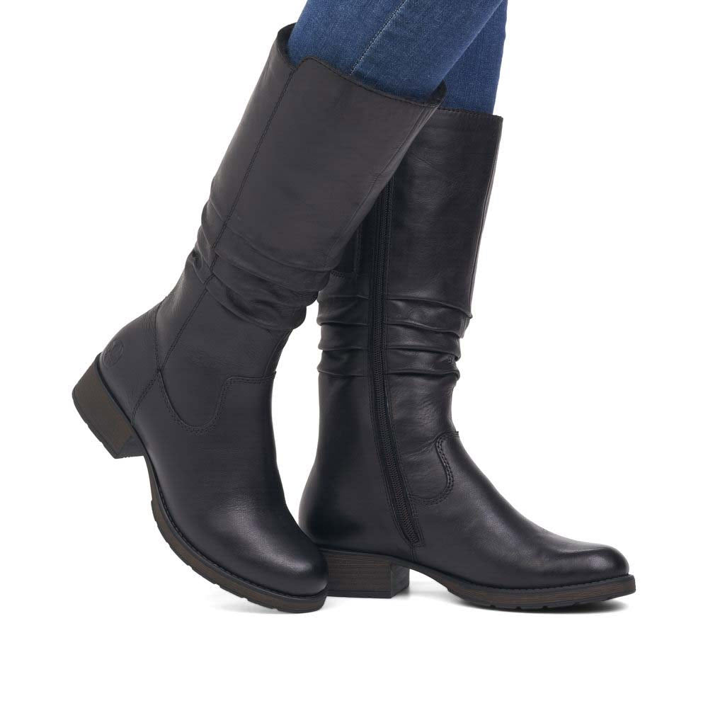 A person wearing Rieker black mid-calf boots and blue jeans, shown from the calves down, against a white background.