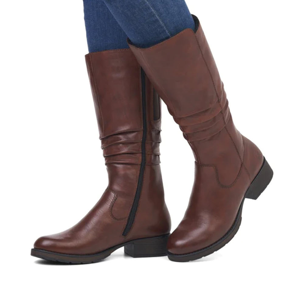A person wearing blue jeans and tall brown Rieker RUCHED MID BOOTIE CHESTNUT mid-calf boots made of breathable leather.