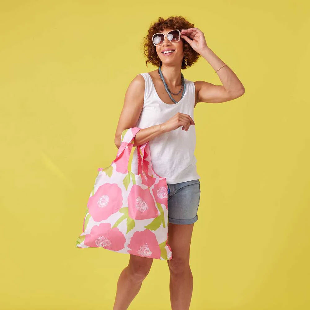 A woman with curly hair, in a white tank top and denim shorts, adjusting her sunglasses and carrying an eco-friendly Rockflowerpaper BLU BAG POPPIES PINK, stands against a yellow background.