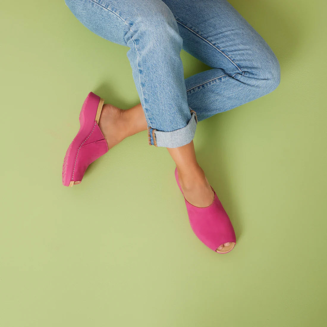 Person wearing Dansko Ravyn Fuchsia peep-toe sandals and blue jeans against a green background, with crossed legs suspended in the air.