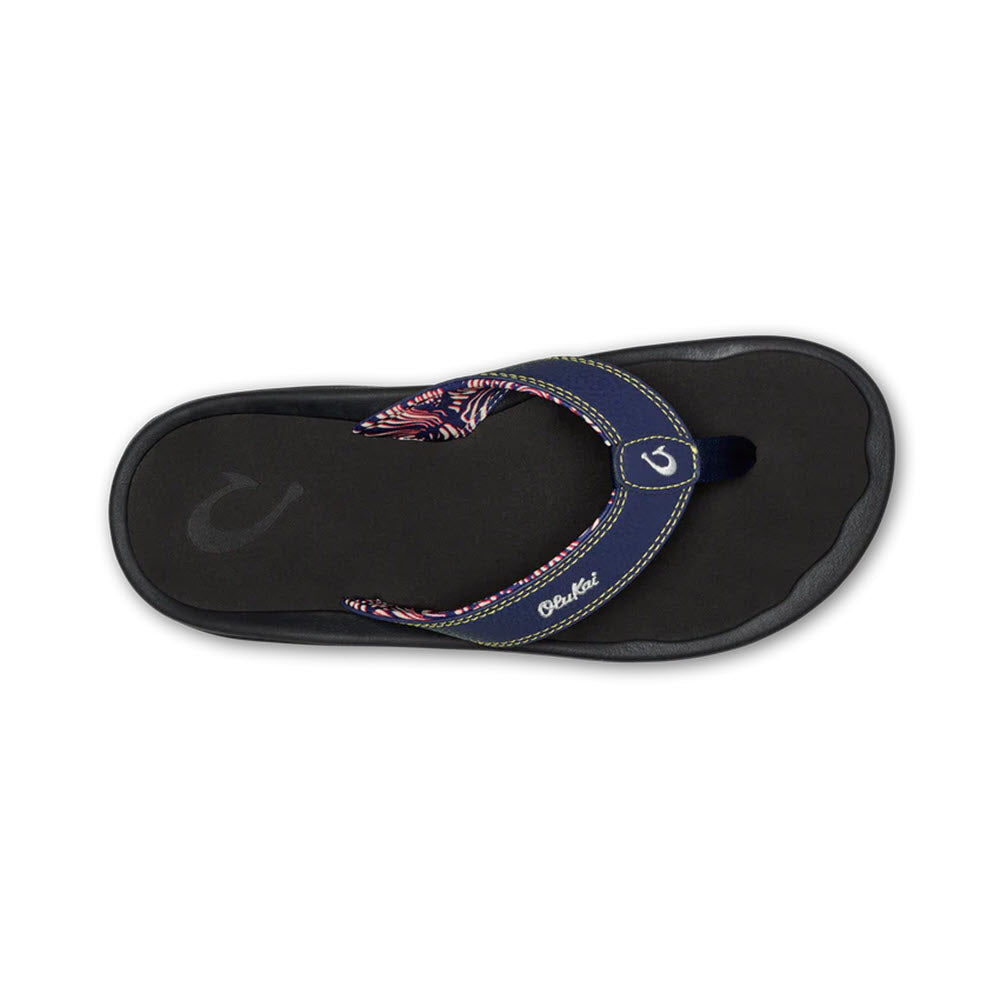 A single Olukai Ohana Flip Flop Navy/Onyx sandal with a black non-slip EVA footbed featuring a patriotic strap design in red, white, and blue, with a logo visible on the heel.