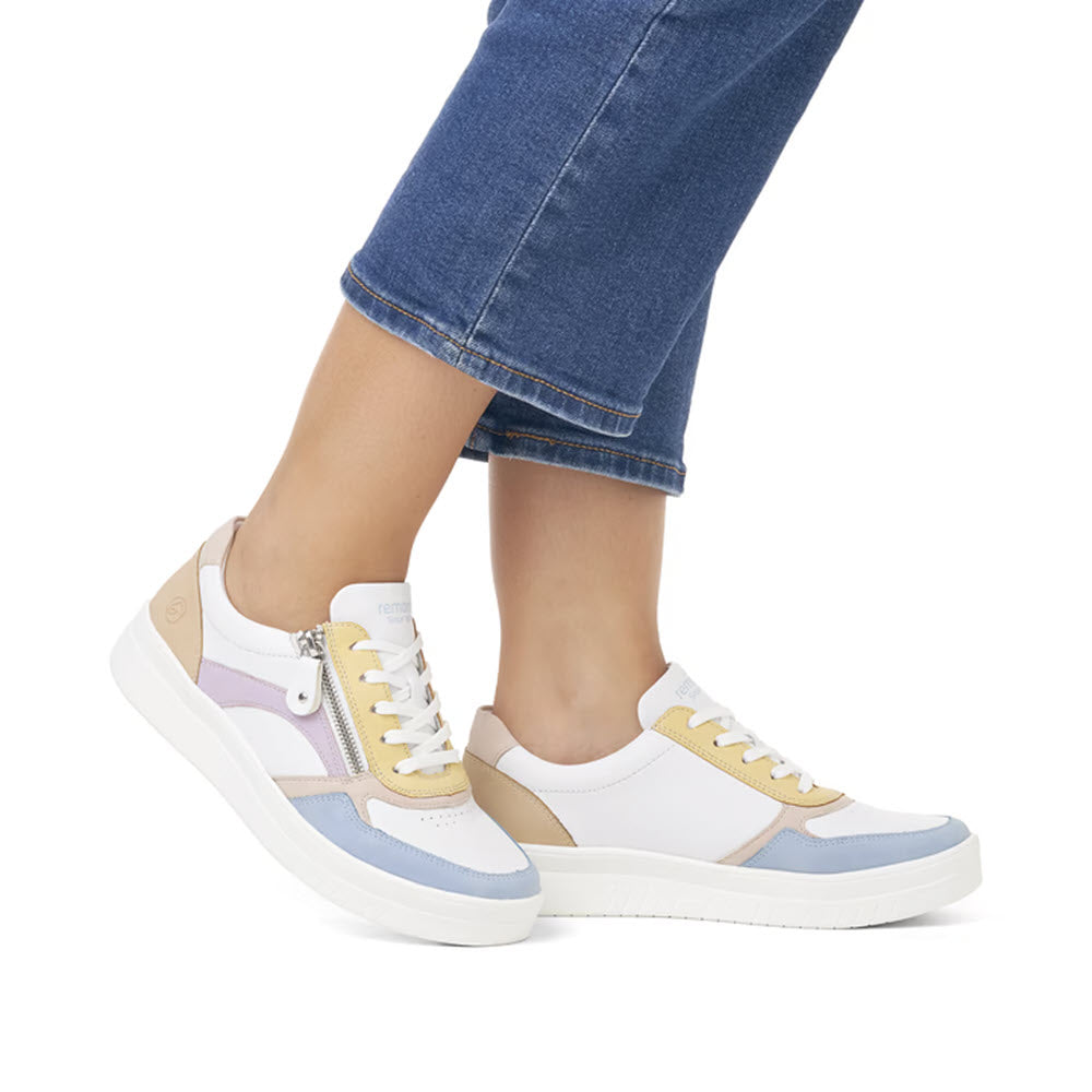 A person wearing white Remonte REMONTE EURO COURT SNEAKER PASTEL MULTI - WOMENS with blue, tan, and lavender accents made from smooth leather, paired with cropped blue jeans.