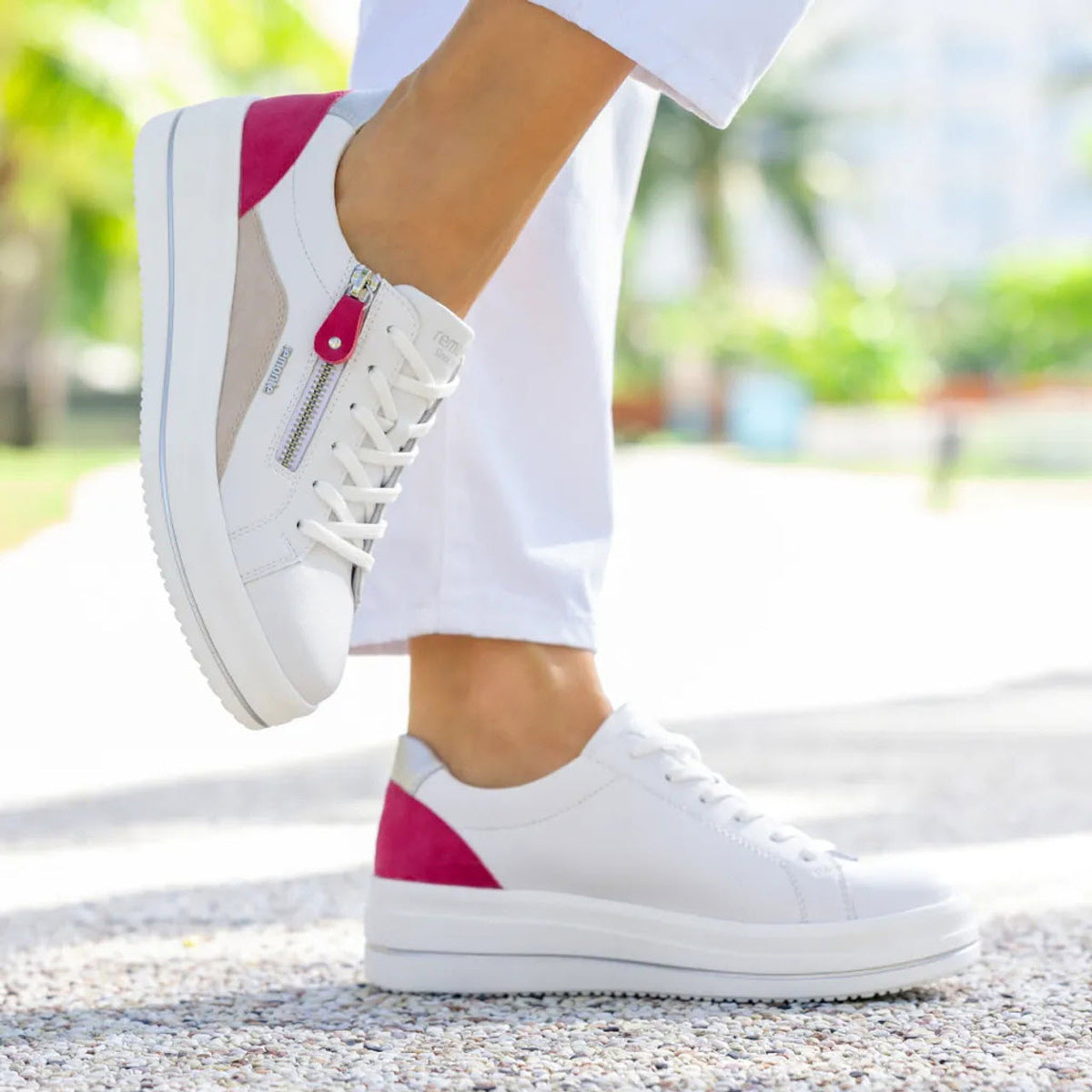 A person wearing white sneakers with pink accents, possibly the Remonte REMONTE EURO COURT SNEAKER FUCHSIA MULTI - WOMENS, and white pants stands and poses outdoors on a sunny day.
