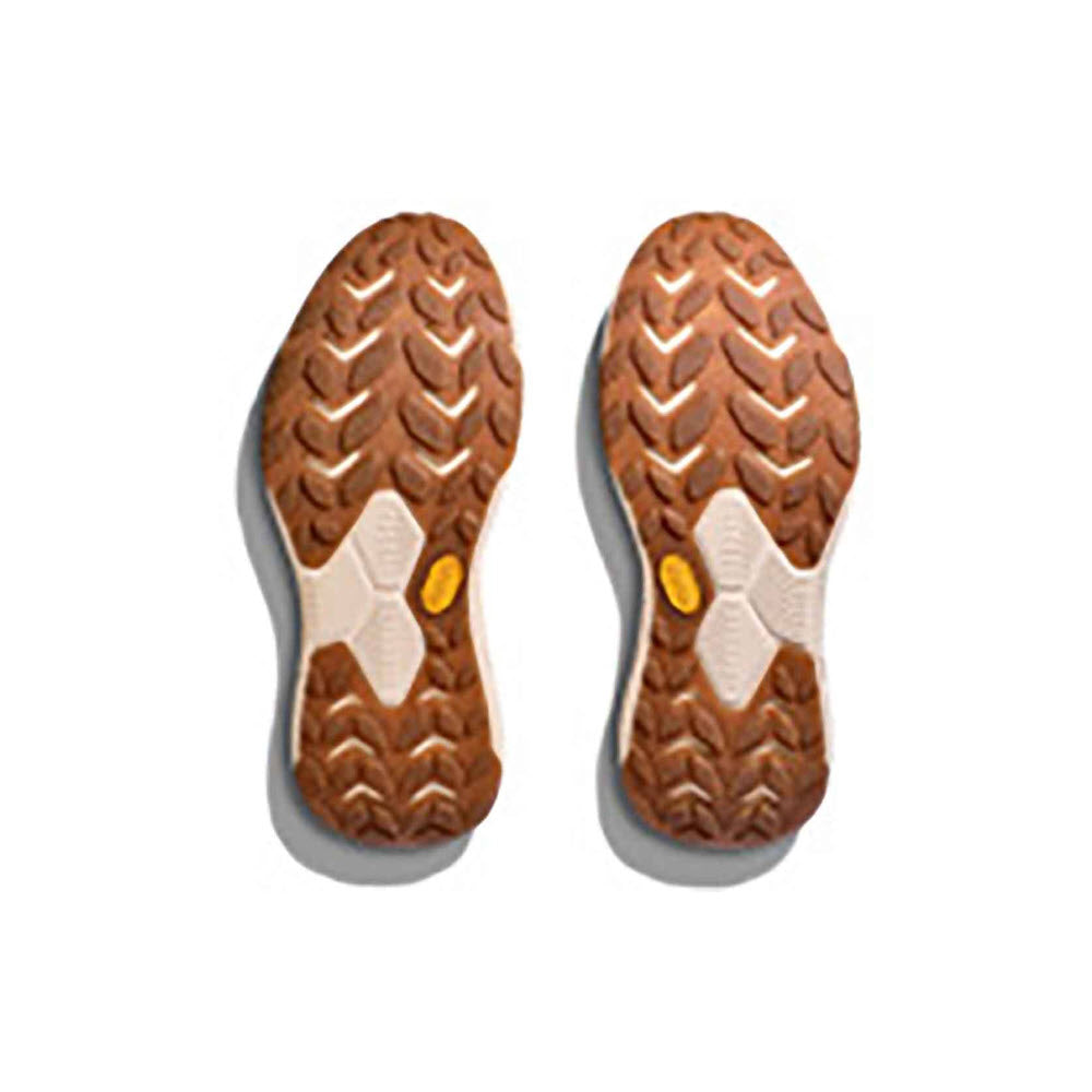 A pair of brown hiking boot soles with rugged tread patterns and white and yellow accents, featuring a Hoka Vibram® EcoStep Natural outsole, isolated on a white background.