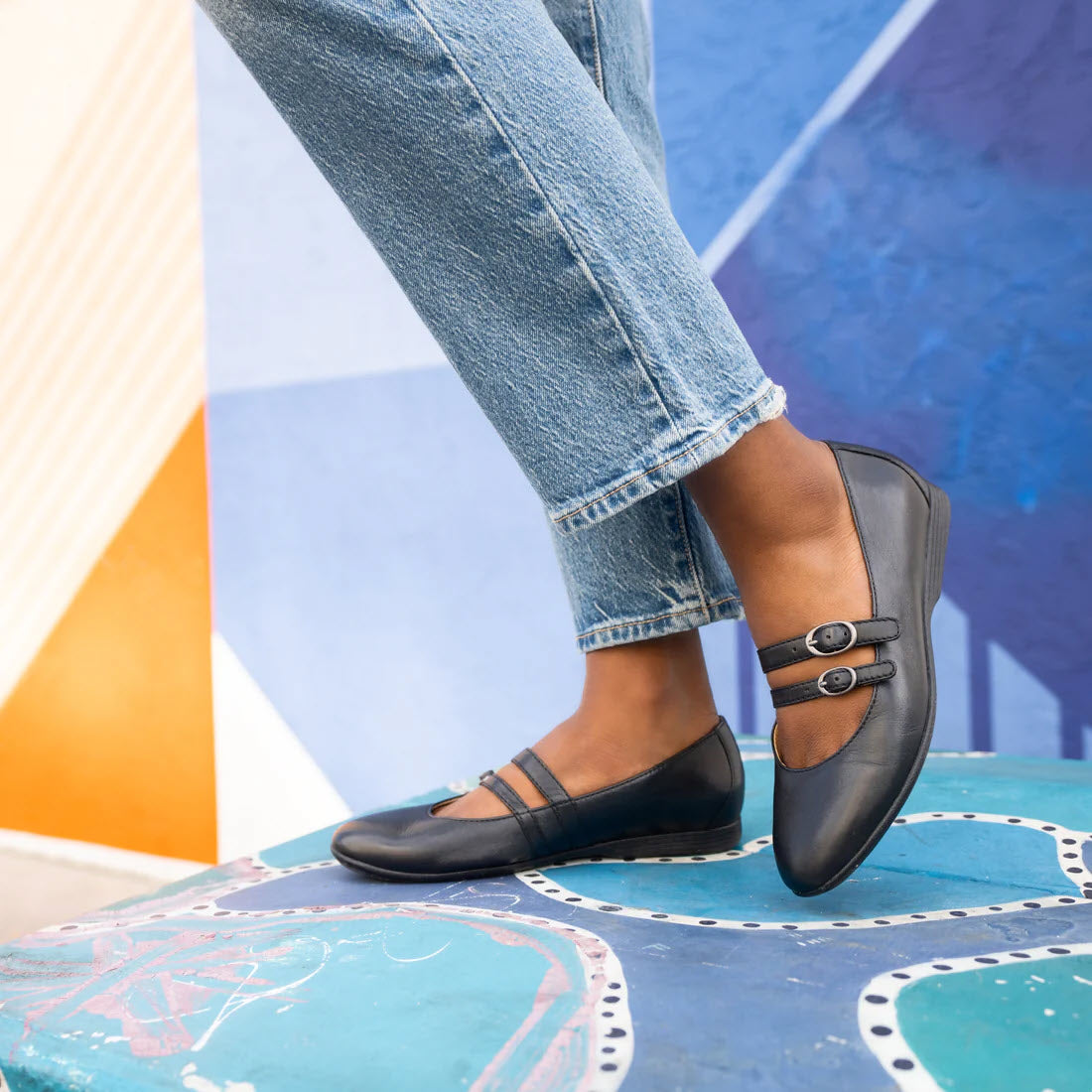 A person standing on a colorful, patterned surface wears cuffed denim jeans and elegant Dansko Leeza Black ballerina flats.