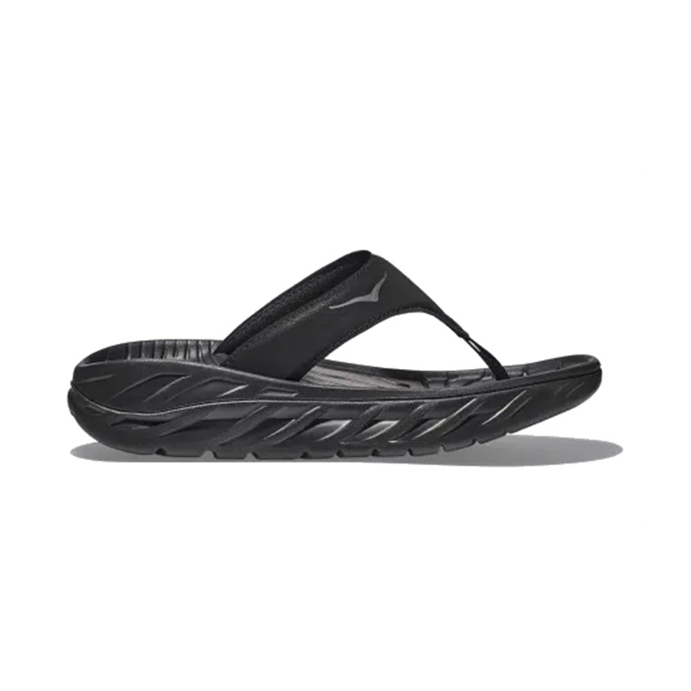 A black sporty Hoka Ora Recovery Flip with an oversized midsole, a thick, textured sole, and a wide over-the-foot strap, on a white background.