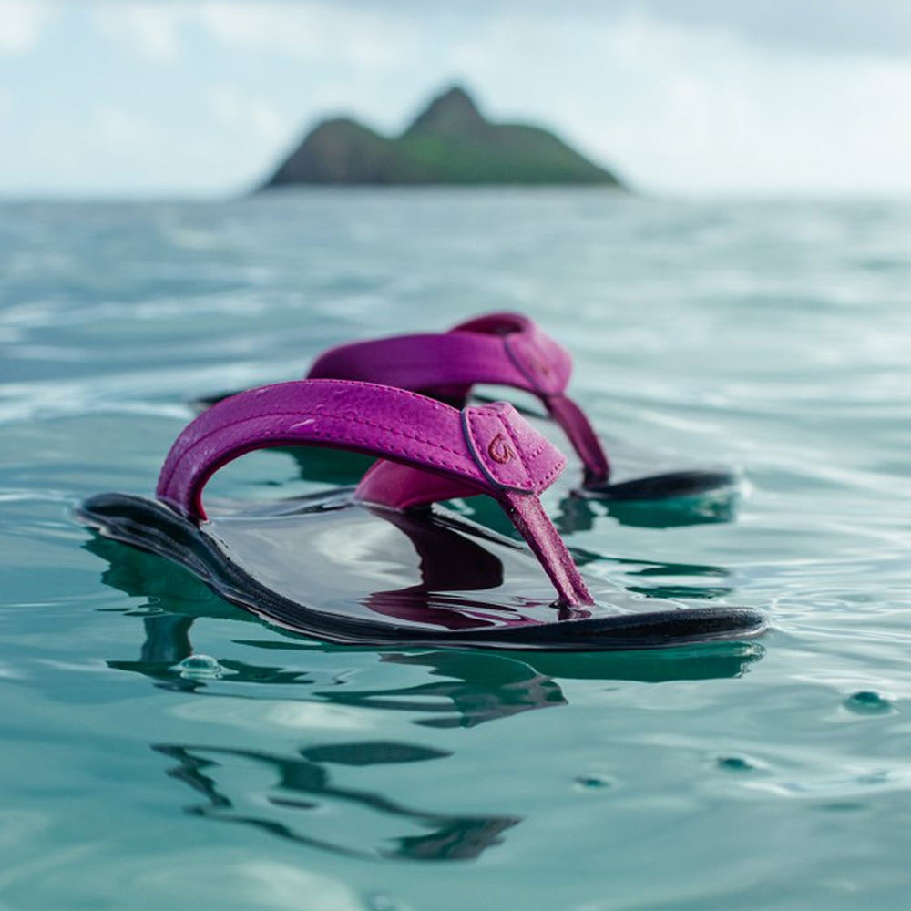 A pair of Olukai Ohana Orchid Flower women&#39;s sandals floating on clear sea water, with a lush green island visible in the background.
