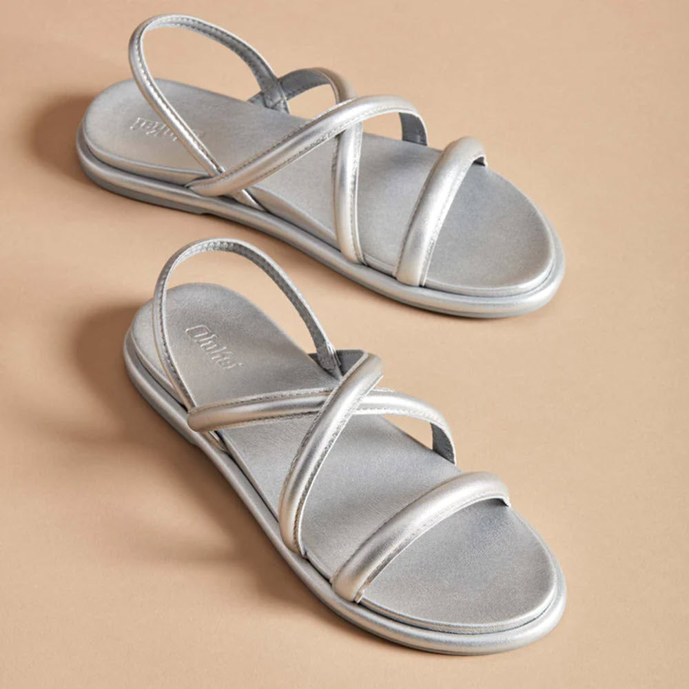 A pair of silver Olukai Tiare Strappy sandals displayed on a beige background.