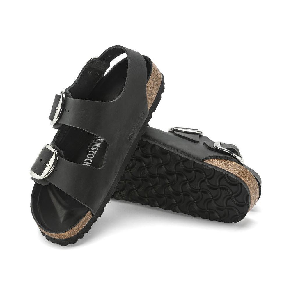 A pair of black Birkenstock Milano Big Buckle Black sandals, featuring a cork footbed and a ridged sole, angled on a white background.