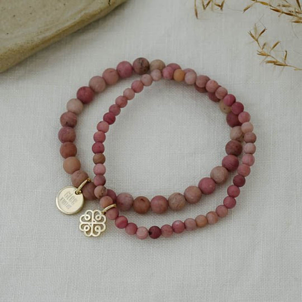 A Glee beaded bracelet in shades of pink and burgundy, displayed on a linen background, with a hypoallergenic gold charm and a stamped tag attached.
