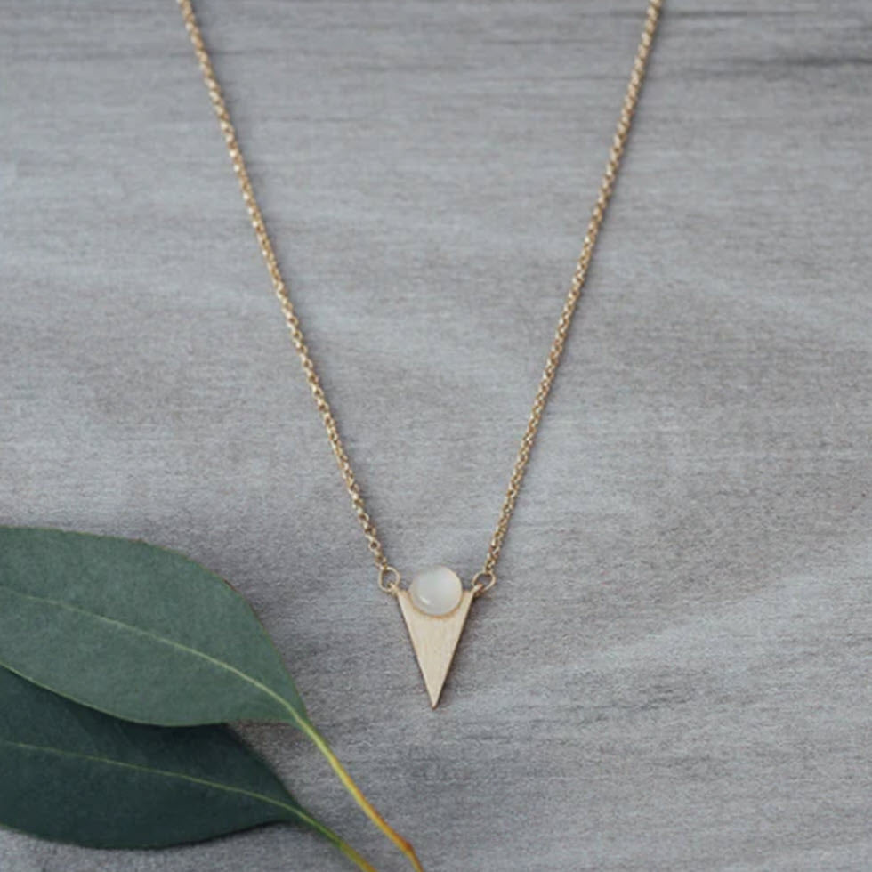 Glee Moon Child necklace in gold with a triangular pendant and a small pearl, displayed on a gray fabric background beside a green leaf. This piece is nickel and lead free.