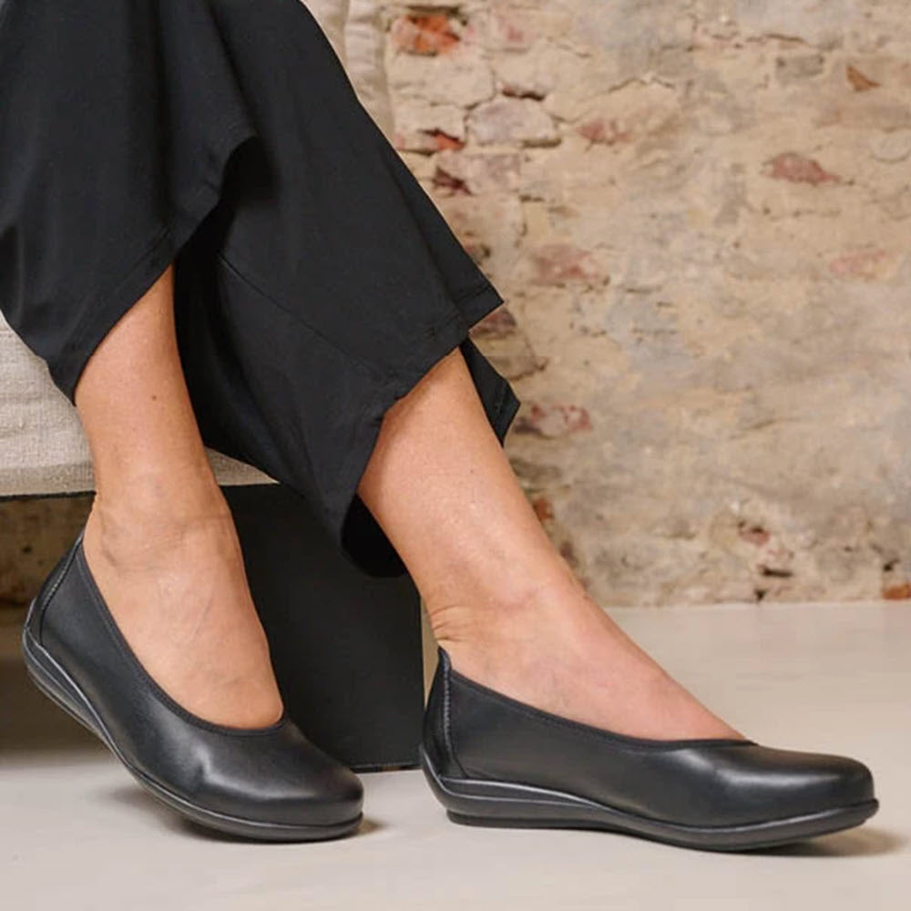 Black leather Wolky Duncan F2F flats with a flexible rubber sole on a woman seated with crossed ankles, showing a brick wall background.