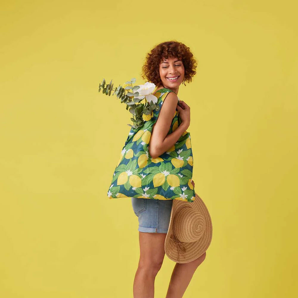 A woman with curly hair, smiling, holding a Rockflowerpaper BLU BAG VINTAGE LEMONS and a hat, standing against a yellow background.