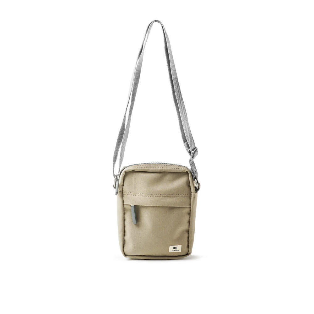 A small, khaki-colored Ori London Bond A Crossbody Coriander with a silver strap designed as a lightweight urban accessory, standing upright against a white background.