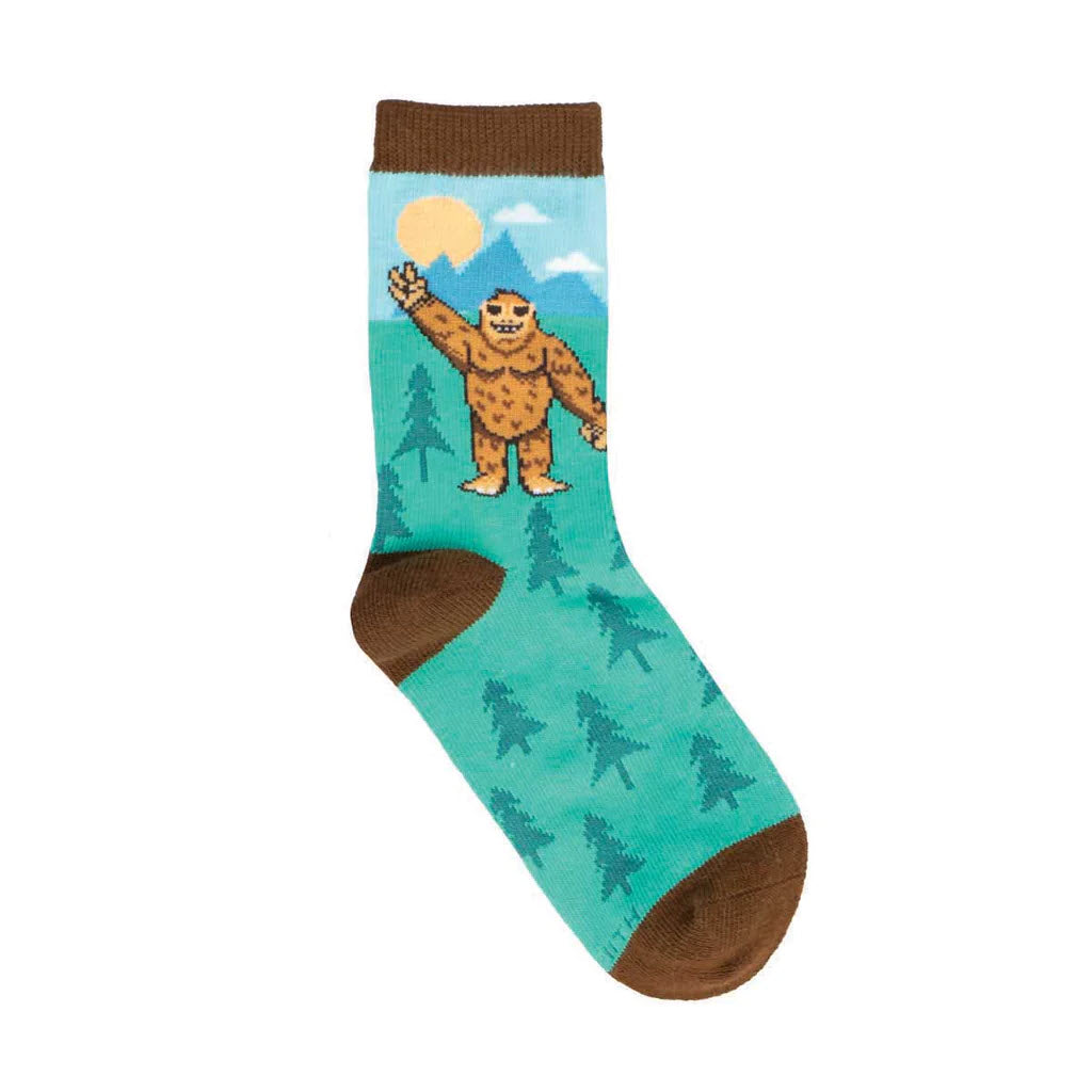 A SOCKSMITH PEACE OUT BIGFOOT CREW SOCKS BLUE - KIDS featuring a cartoon-style bigfoot, a mythical creature, holding a glowing sun, with mountains and trees in the background, and brown heel and toe patches.