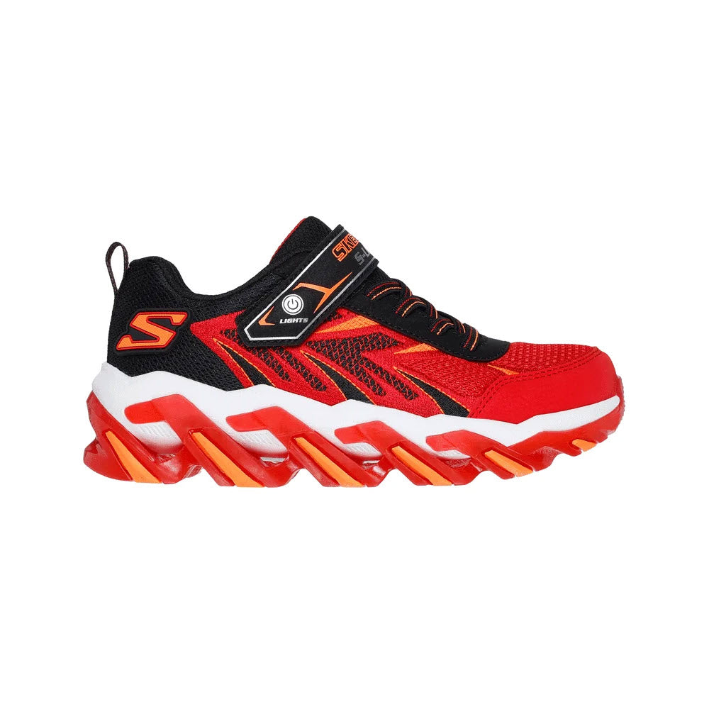 A bright red and black athletic sneaker with orange accents, a chunky sole, a Velcro strap, mesh panels, and the letter S logo on the side. The shoe features a futuristic and sporty design.