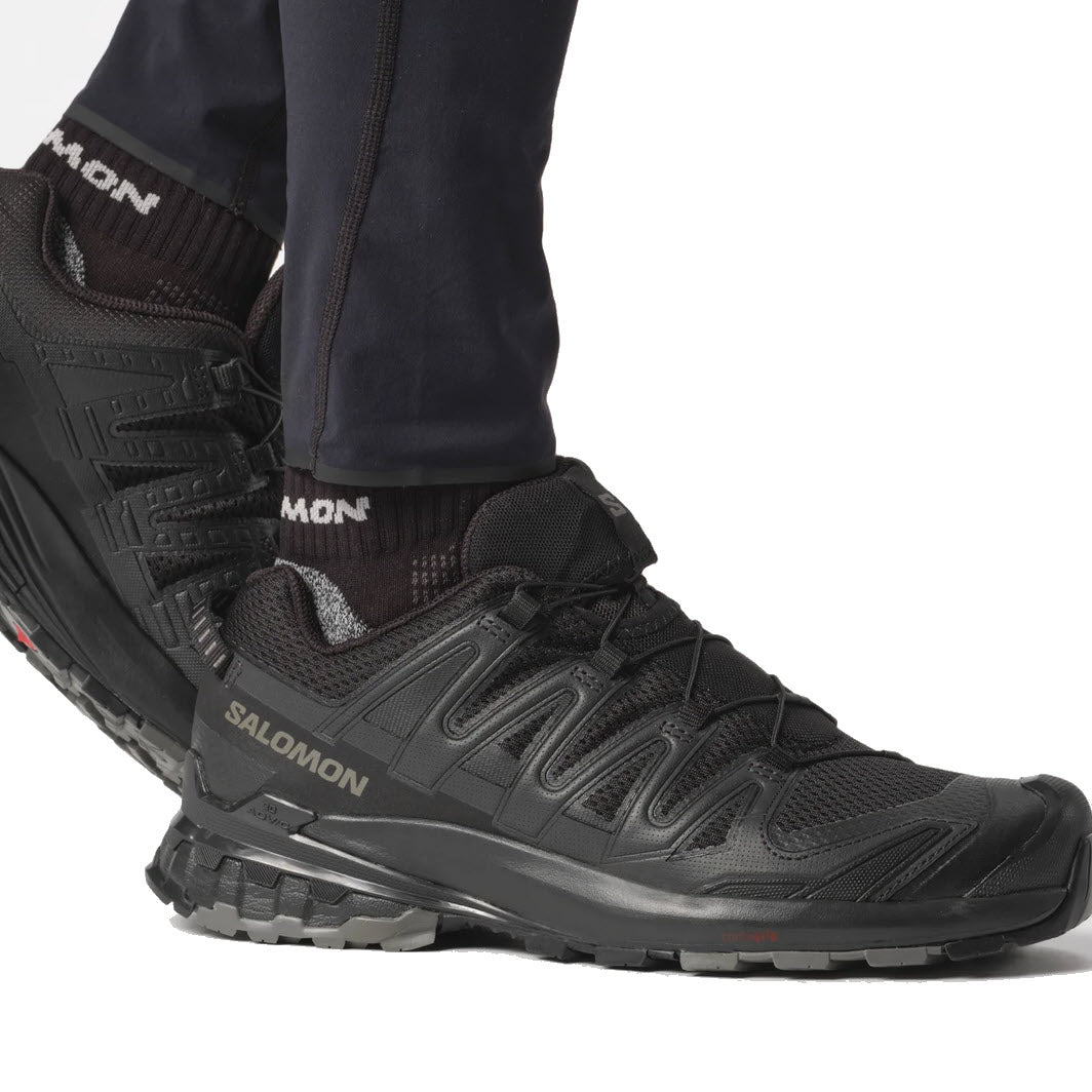 A close-up of a person&#39;s feet wearing the black Salomon XA PRO 3D V9 hiking shoes and navy pants, focused on the shoes&#39; intricate Contagrip tread design.