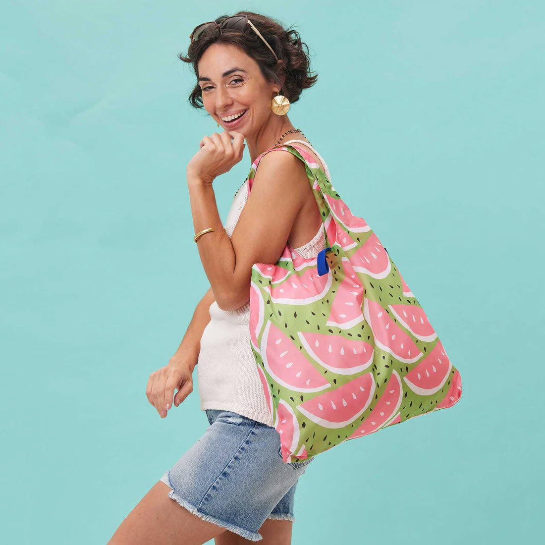 Woman smiling, carrying a large BLU BAG WATERMELON reusable shopping bag on her shoulder, dressed in a white top and denim shorts, against a teal background.