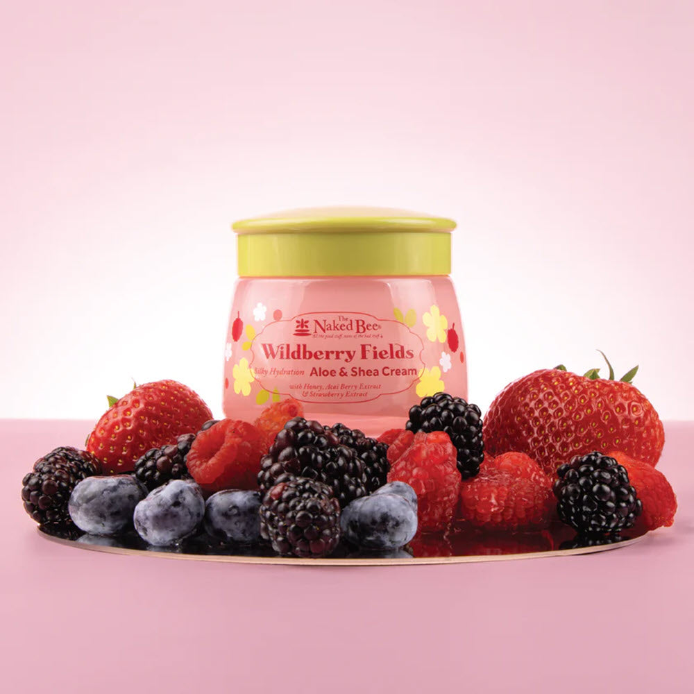 A jar of The Naked Bee Wildberry Fields cream sits on a surface surrounded by fresh strawberries, blueberries, blackberries, and raspberries, all set against a soft pink background.